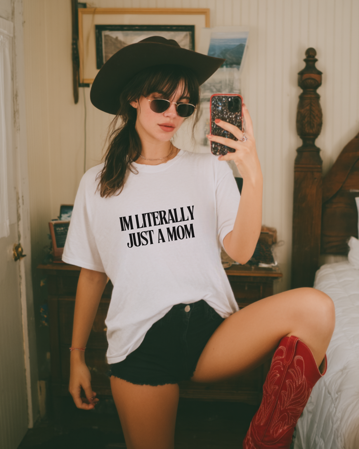 Woman taking a mirror selfie wearing a white t-shirt with text, black hat, sunglasses, and red boots in a bedroom.