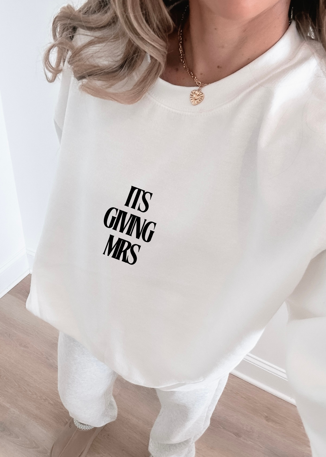 White sweatshirt with 'ITS GIVING MRS' text worn by a person indoors.