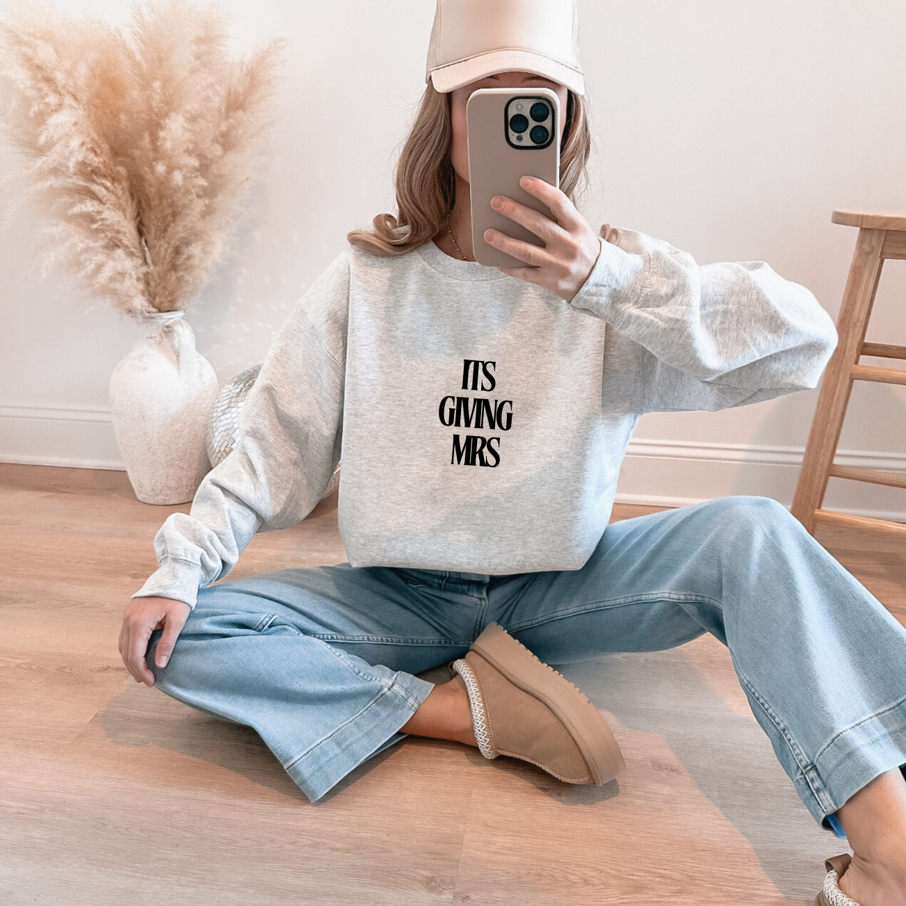 Person wearing a sweatshirt with text (It's Giving Mrs), sitting on the floor in a home setting.