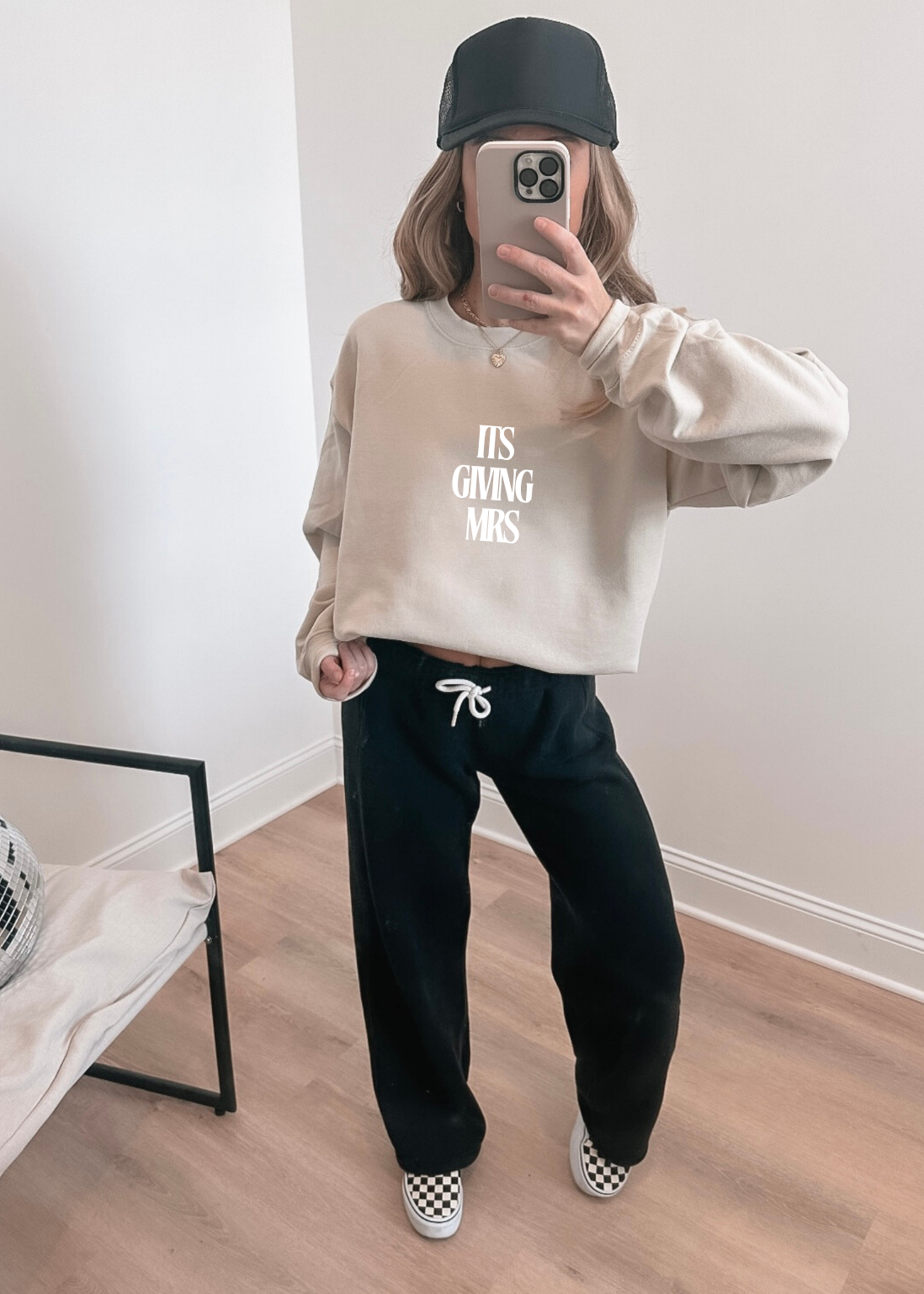 Person wearing a beige sweatshirt with text (it's giving Mrs) and black pants in a room.