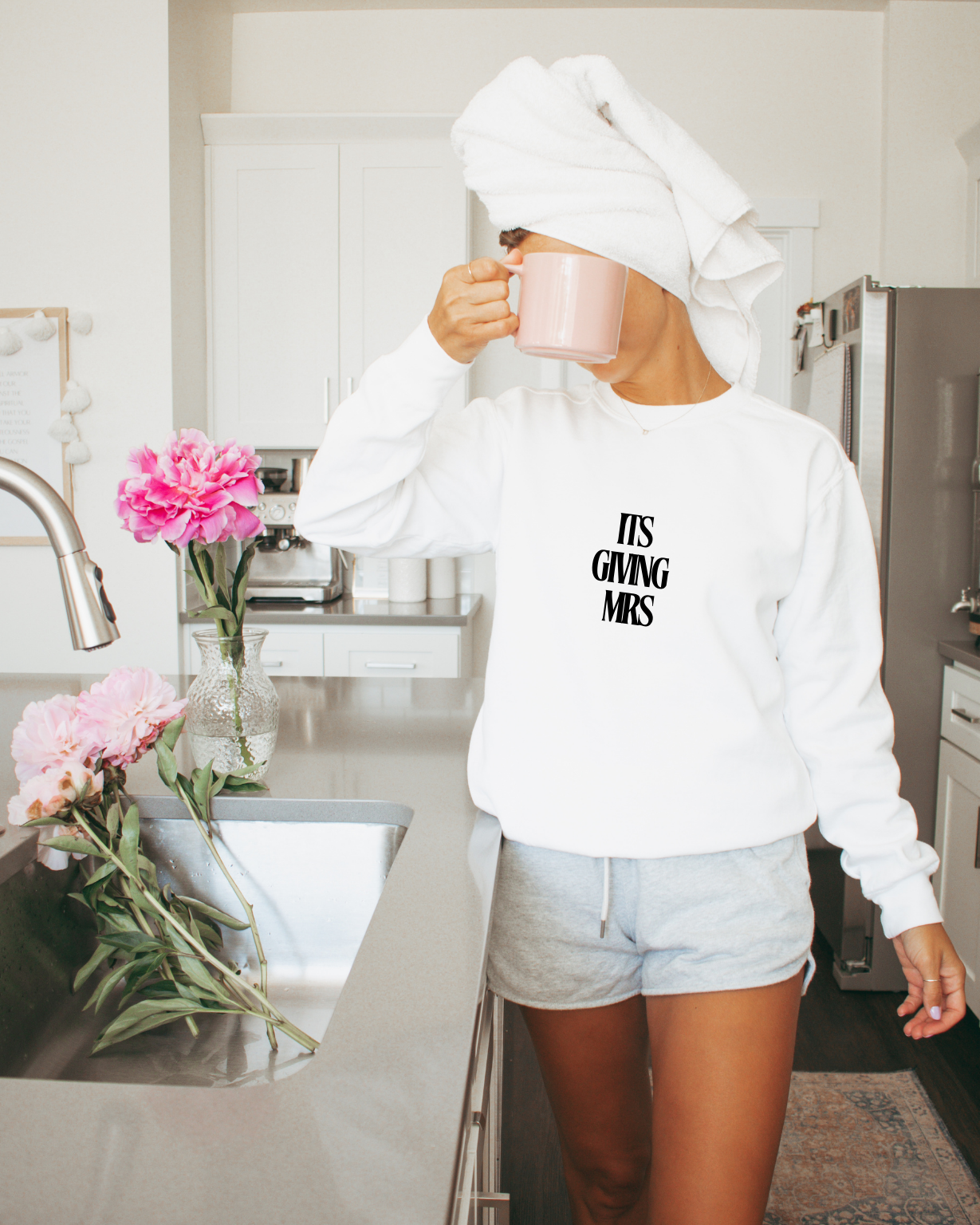 Person in a kitchen wearing a white sweatshirt with ITS GIVING MRS, holding a mug.