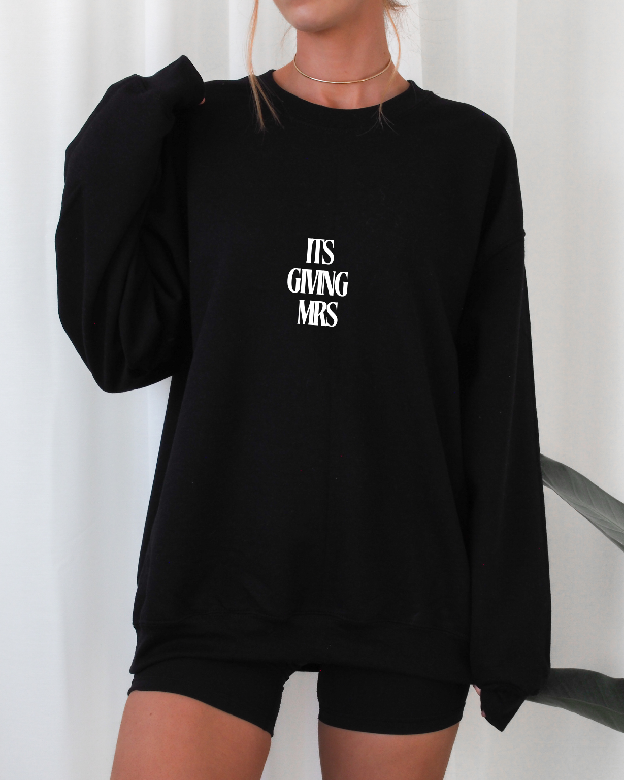 Person wearing a black sweatshirt with text (ITS GIVING MRS) on a white background
