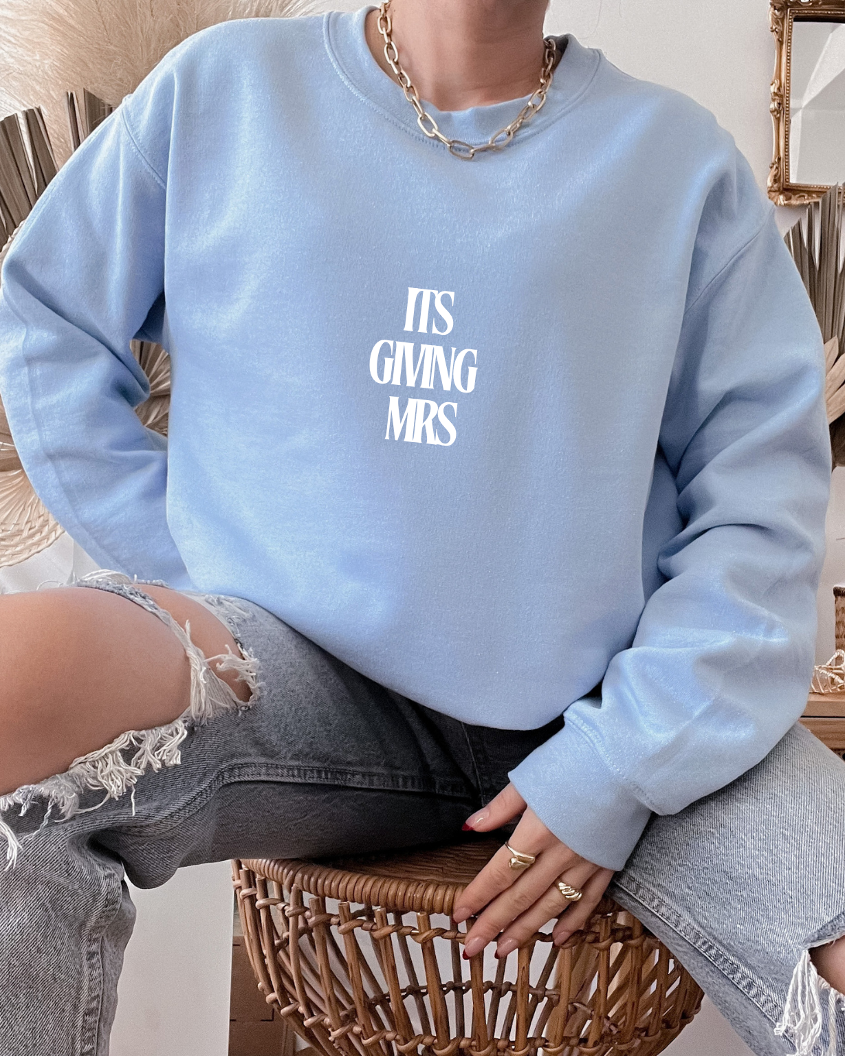 Person wearing a light blue sweatshirt with text (ITS GIVING MRS), sitting on a woven chair.