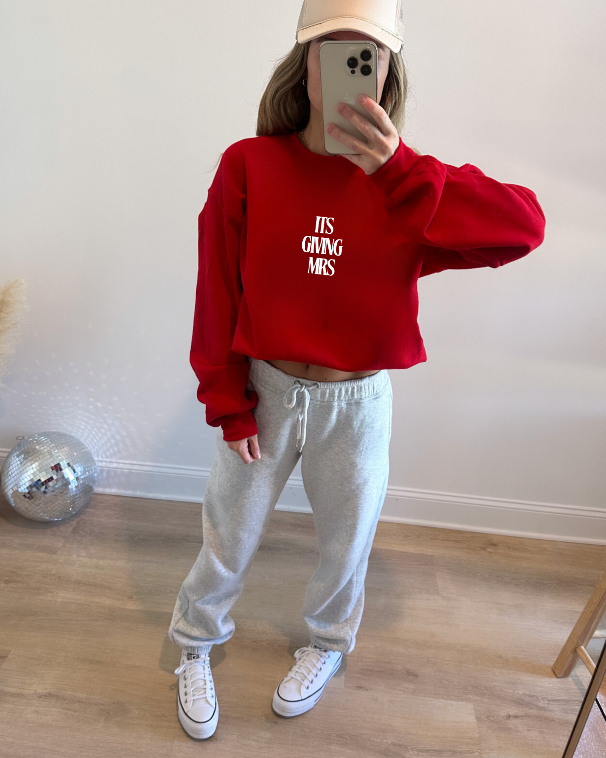 Person wearing a red sweatshirt with text (ITS GIVING MRS) and gray sweatpants in a room.