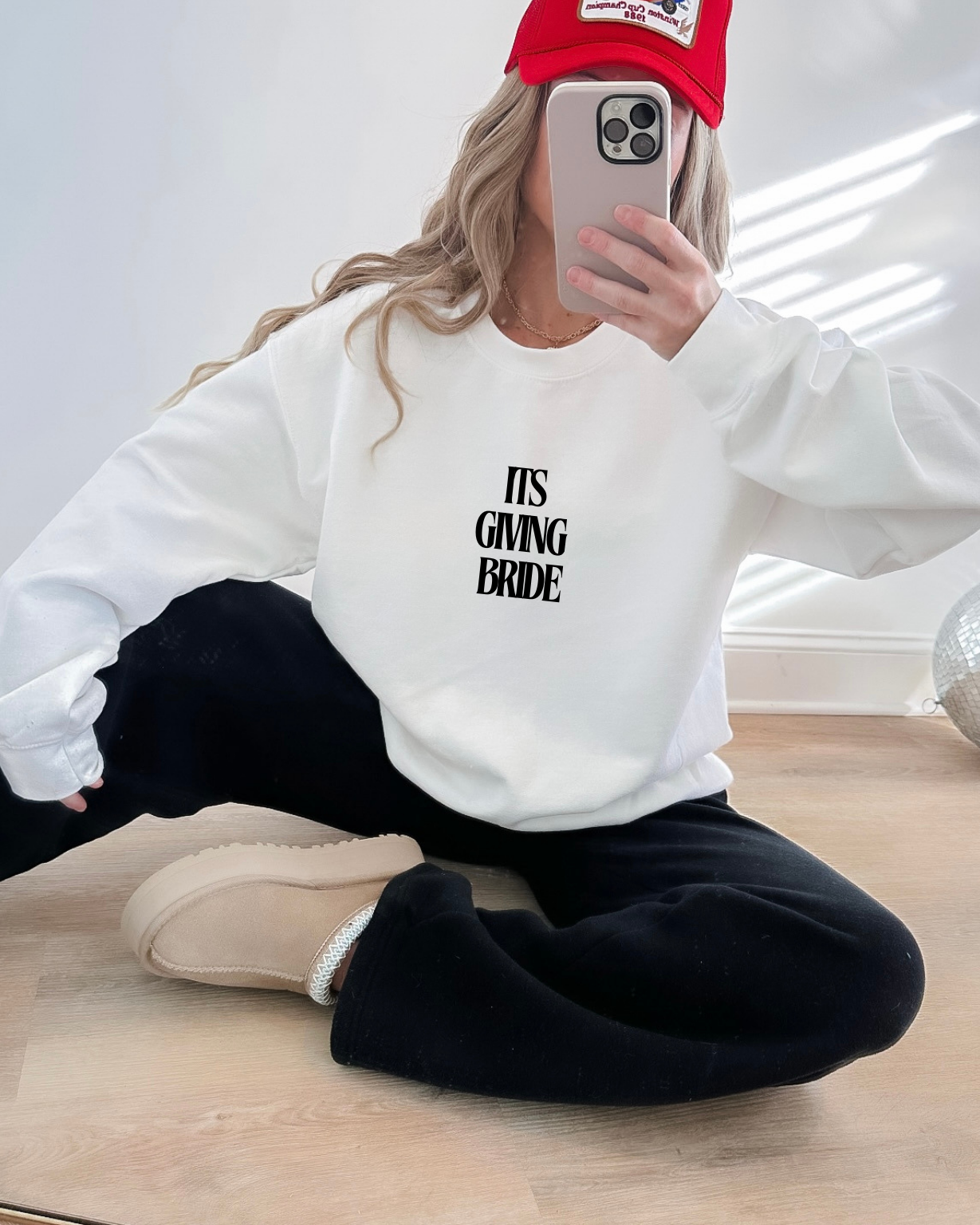 Person wearing a white sweatshirt with 'IT'S GIVING BRIDE' text, sitting on the floor.