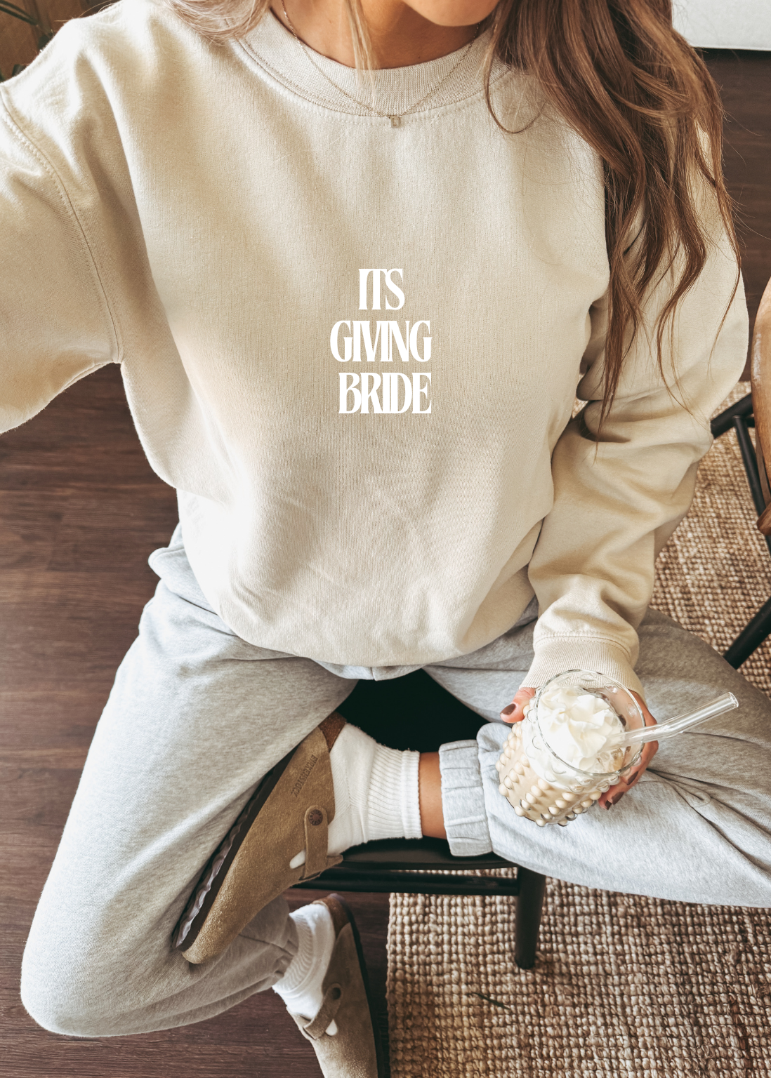 Person wearing a beige sweatshirt with 'IT'S GIVING BRIDE' text, sitting on a chair.