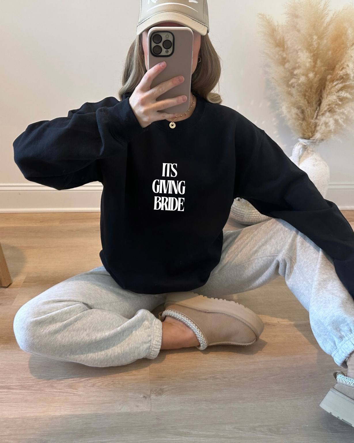 Person wearing a black hoodie with 'IT'S GIVING BRIDE' text, sitting on a wooden floor.