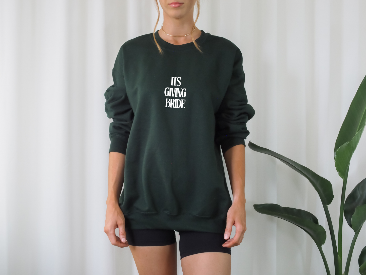 Person wearing a dark green sweatshirt with 'IT'S GIVING BRIDE' text against a white curtain background.