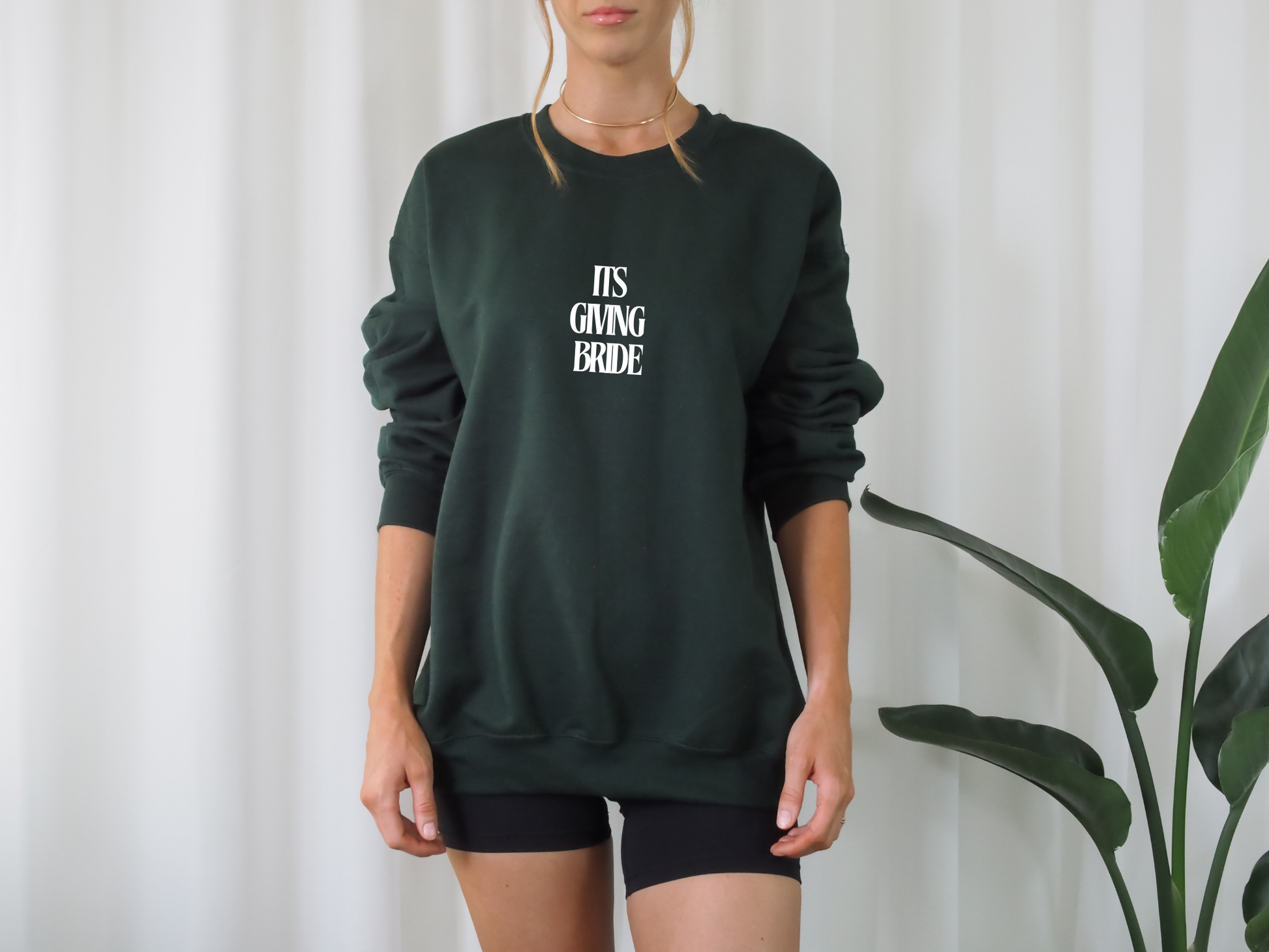Person wearing a dark green sweatshirt with 'IT'S GIVING BRIDE' text against a white curtain background.