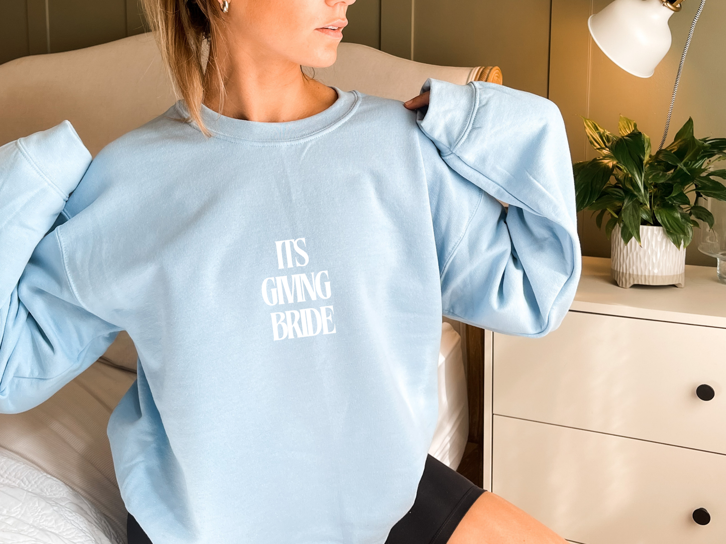 Woman wearing a light blue sweatshirt with 'IT'S GIVING BRIDE' text, sitting on a couch.