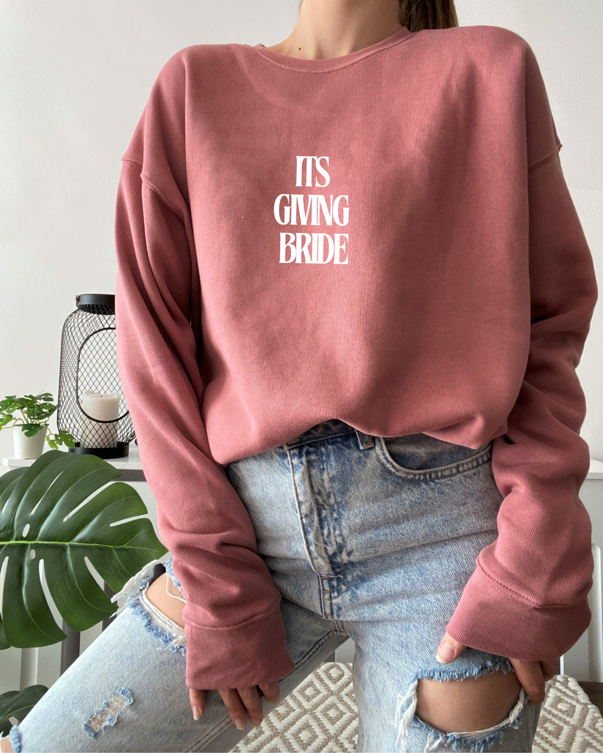 Person wearing a pink sweatshirt with 'IT'S GIVING BRIDE' text, sitting indoors.